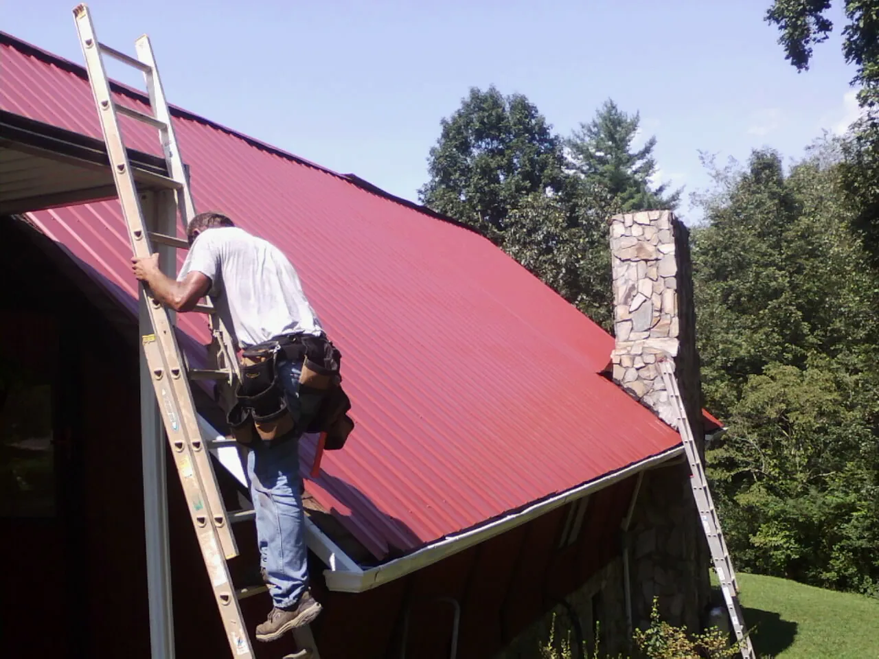 Roofer on a ladder beside a red metal roof and stone chimney — Soffit & Fascia Repair by Roofing Brothers in Van Nuys